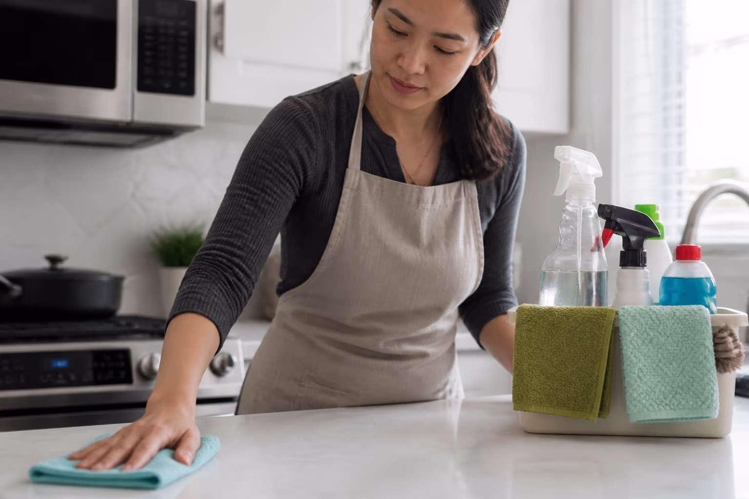 A professional home cleaner in an apron holding a caddy of cloths and cleaning supplies in a bright kitchen, ready to tackle the hidden spots most people miss.