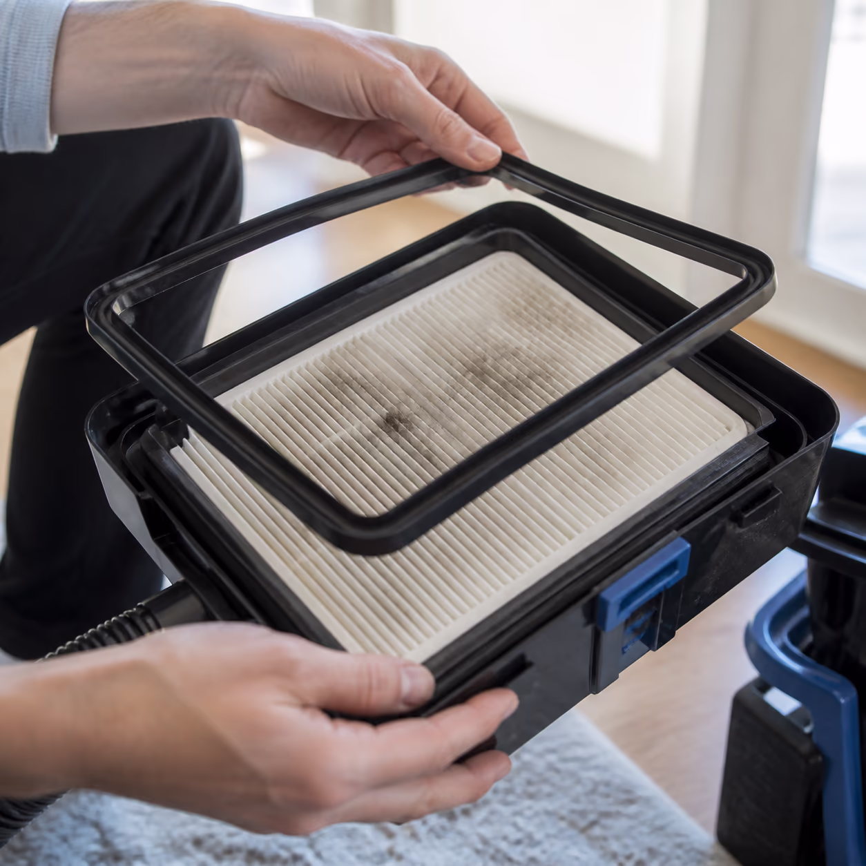 A close view of a sealed HEPA vacuum cleaner filter housing with its rubber gasket seal open, fine grey dust captured on the pleated white filter media in soft window light.