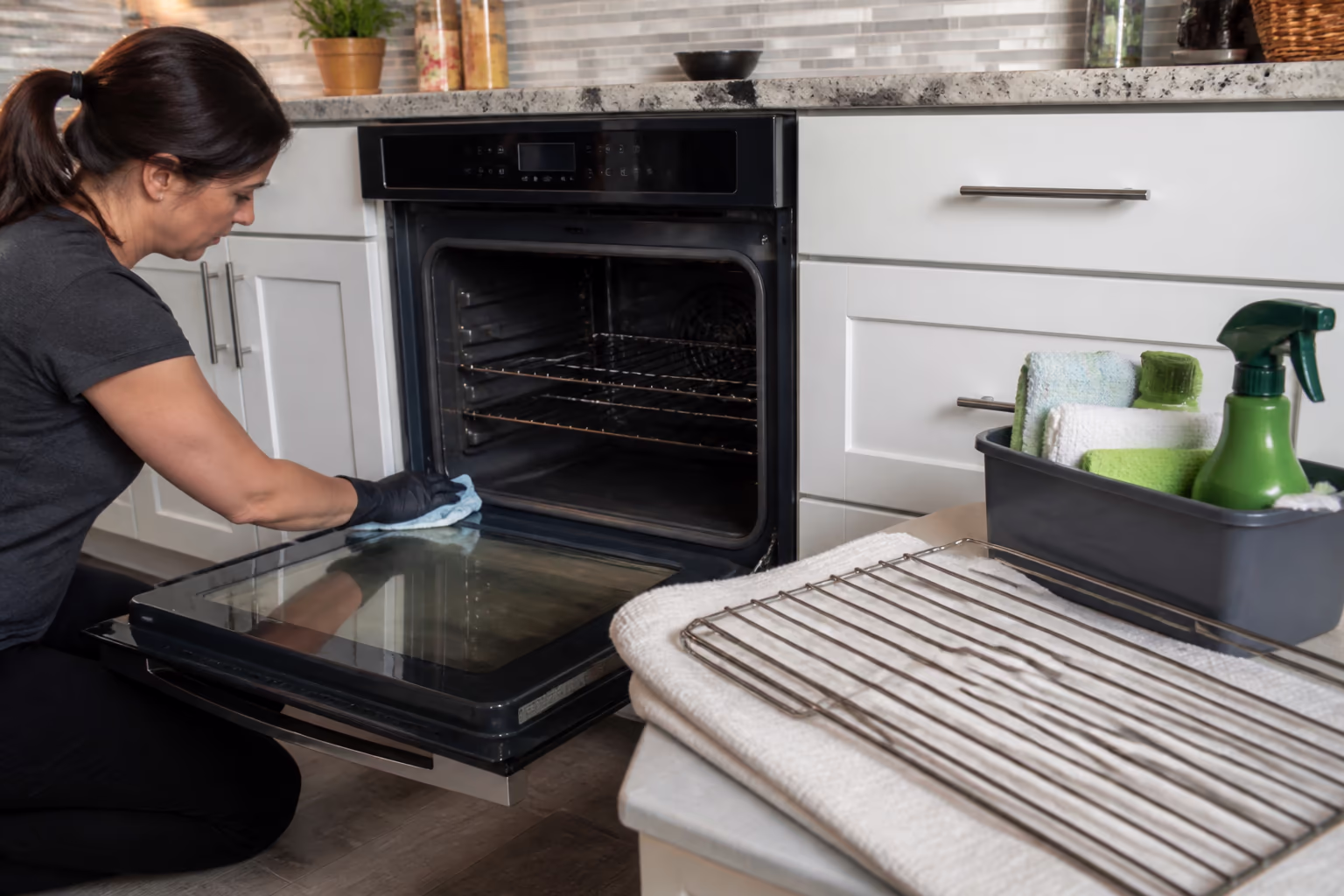 A cleaning technician finishing a kitchen deep-clean, wiping an oven door while a freshly restored oven rack sits on a folded towel beside a caddy of cloths and green bottles.