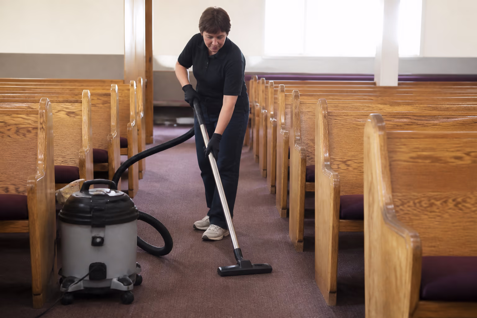 A professional cleaner running a sealed commercial HEPA vacuum down a carpeted sanctuary aisle between rows of wooden pews.
