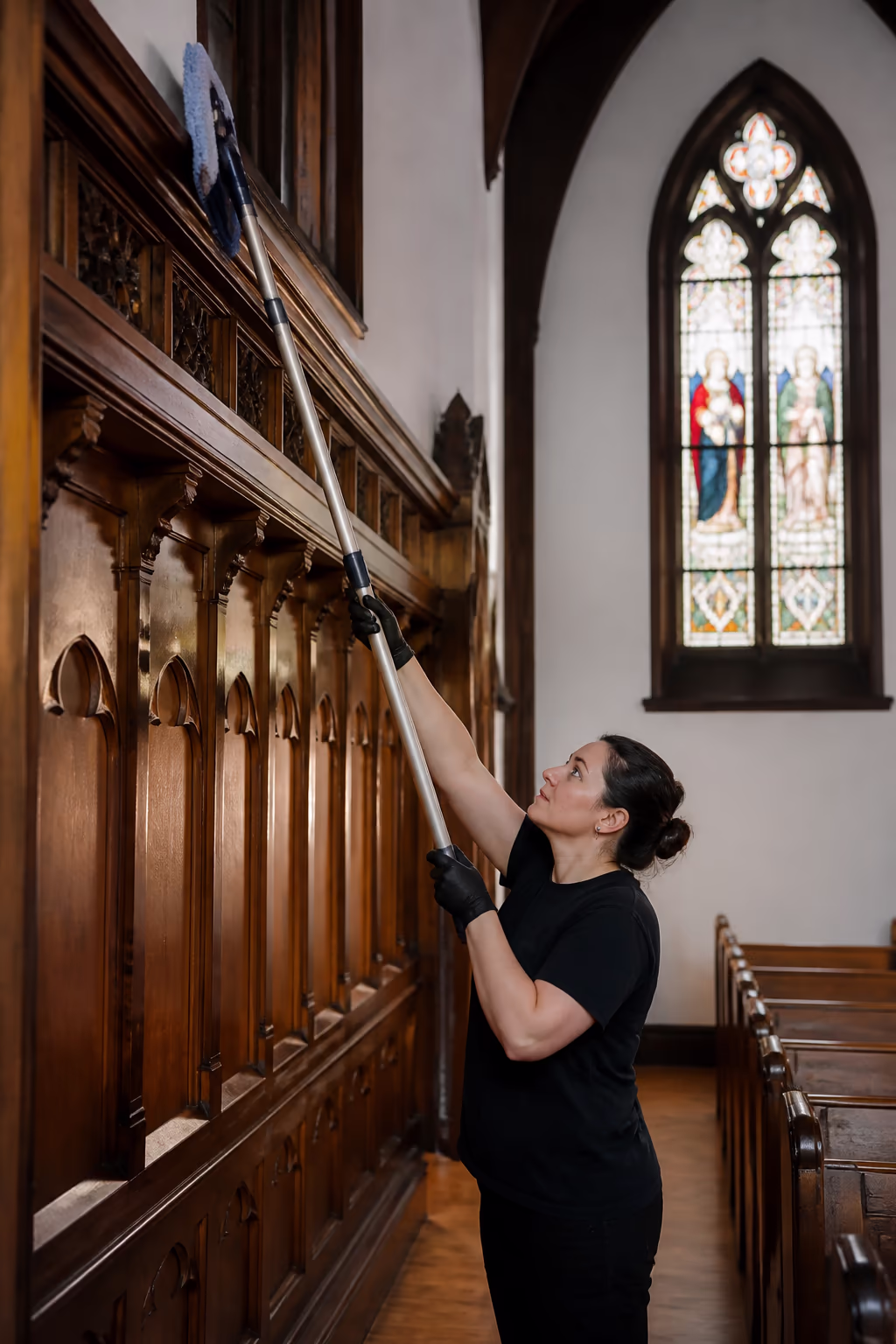 A cleaning technician dusting a historic church sanctuary aisle with a microfibre extension tool between wooden pews, with soft light through a stained glass window.