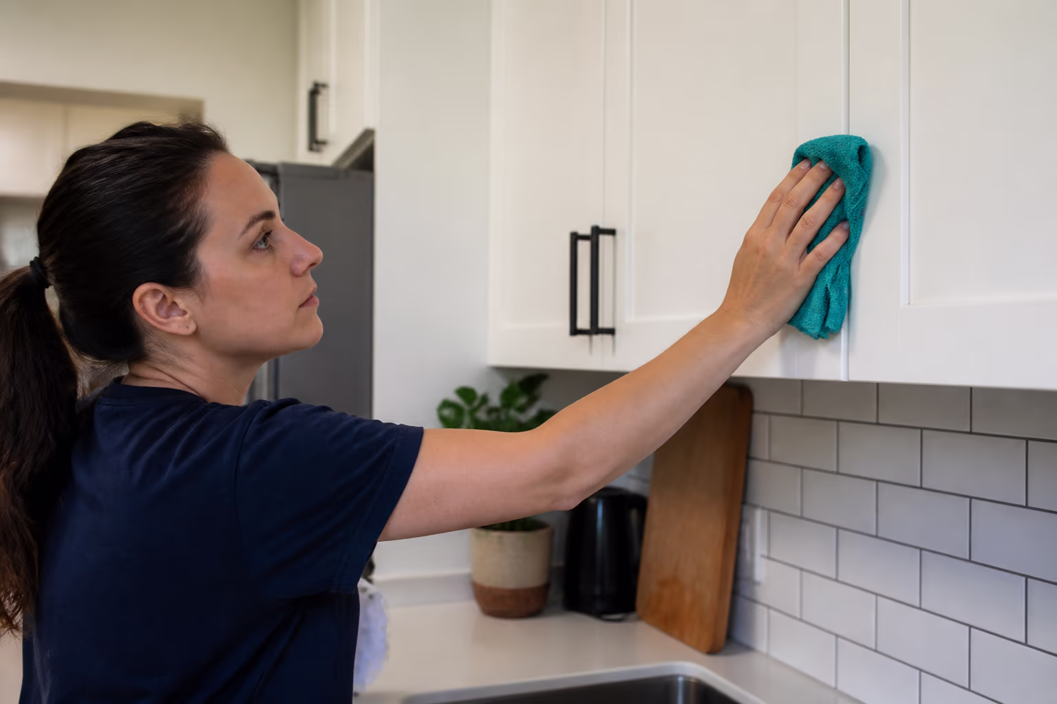 A cleaner in an apron and yellow gloves holding a caddy of cloths and cleaning bottles in a bright kitchen, ready to degrease cabinet fronts.