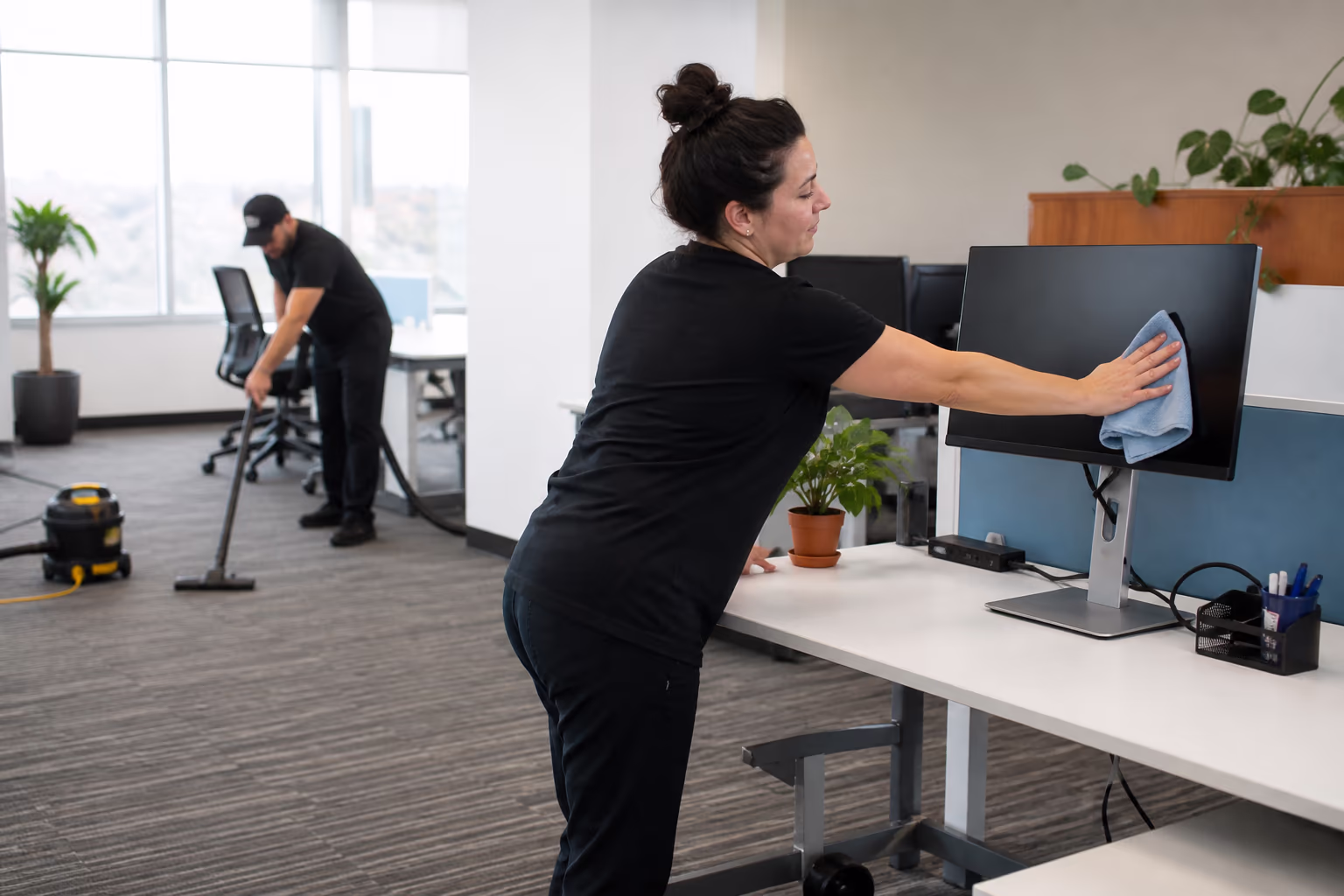 Three cleaners in a bright open-plan office: one vacuuming the floor, one wiping a monitor and one washing a window with a pole.