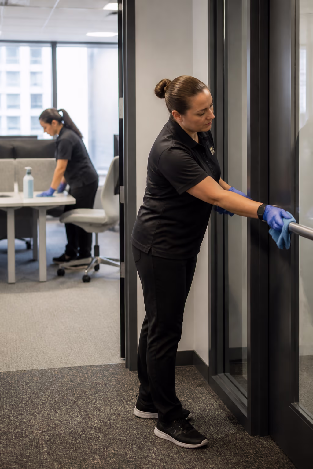 Two cleaners servicing an open-plan office, one detailing a shared desk while the other wipes a door push bar in a bright Ottawa workplace.