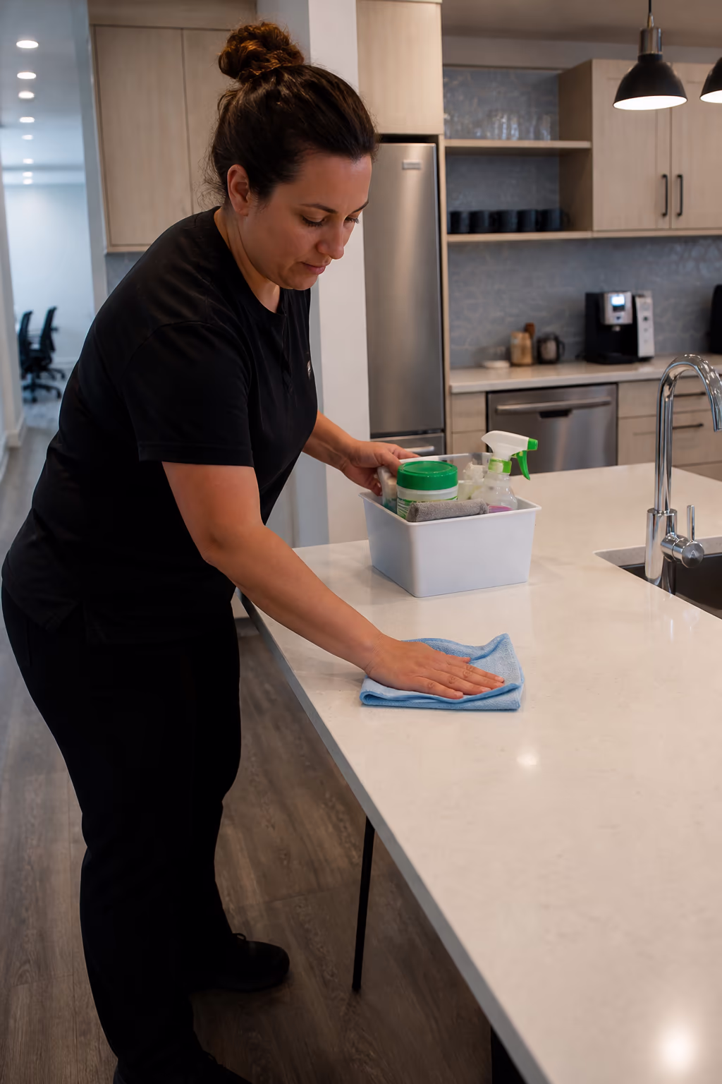A cleaner wiping a kitchen counter and restocking supplies in a modern hybrid office break room between scheduled visits in Ottawa.