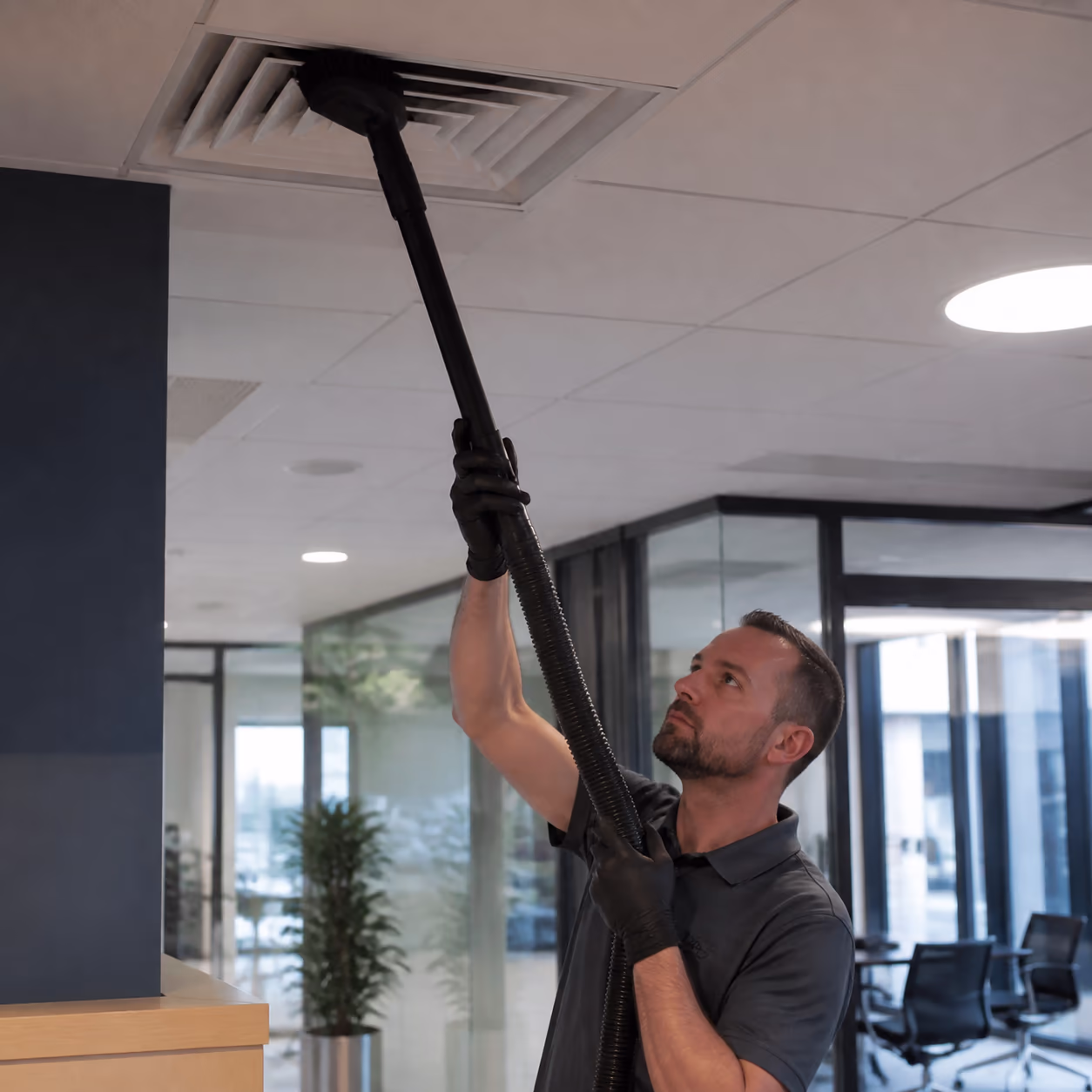 Commercial cleaners in a bright Ottawa office pulling built-up dust from high ceiling HVAC vents and window sills with extendable tools during a periodic deep clean.