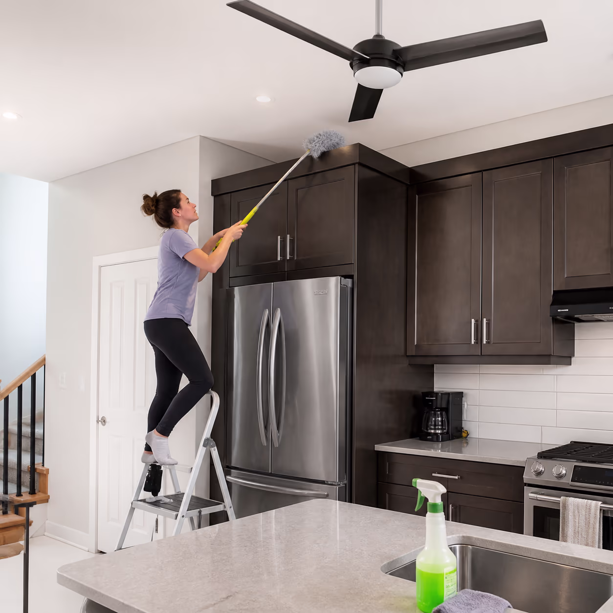 A cleaner on a sturdy step stool dusting the tops of kitchen cabinets and a ceiling fan with an extendable duster in a bright Ottawa home.