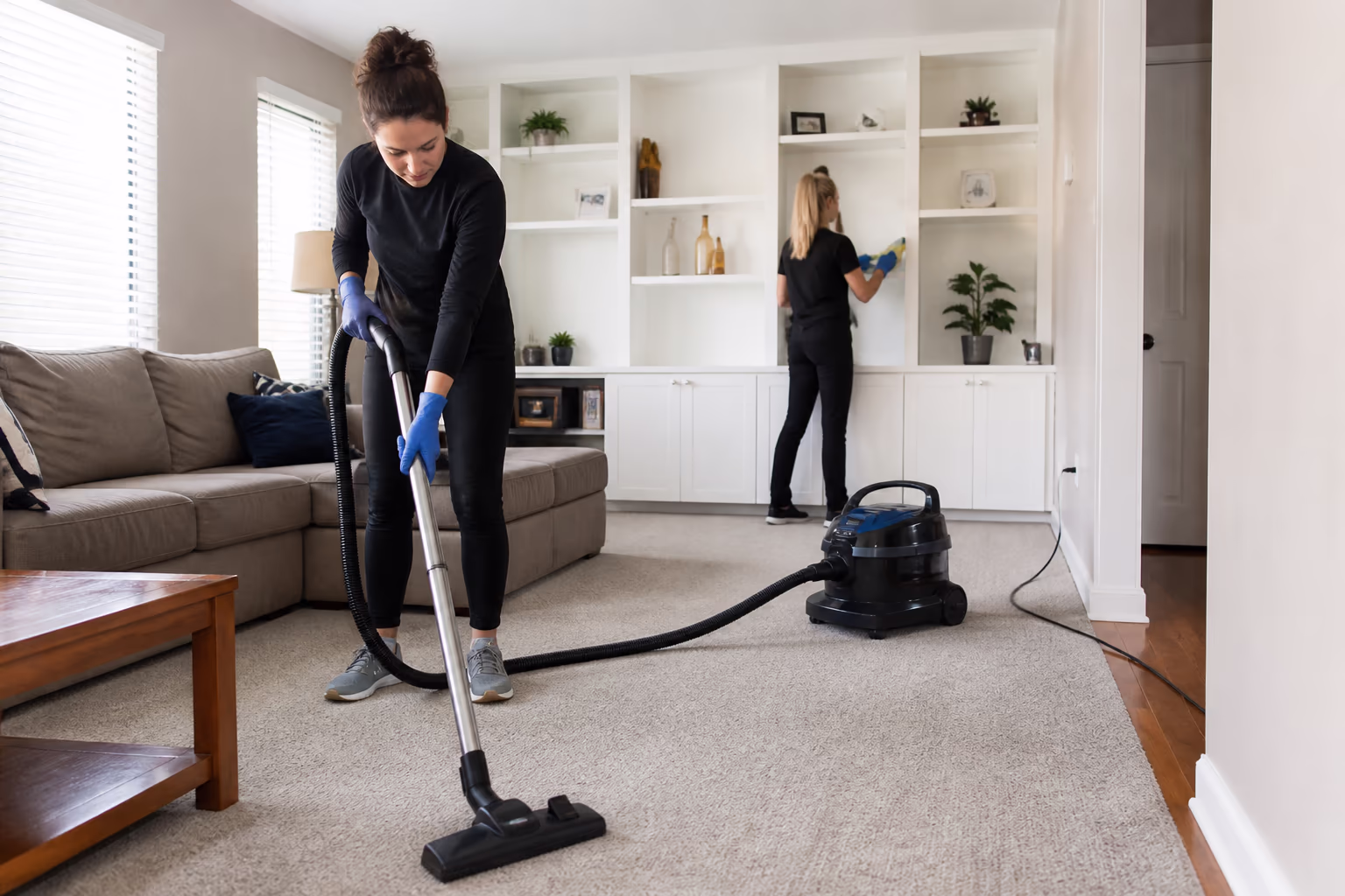 A cleaning technician vacuuming a carpeted floor with a HEPA vacuum and soft brush attachment while a second cleaner dusts a shelf behind, in an Ottawa home.