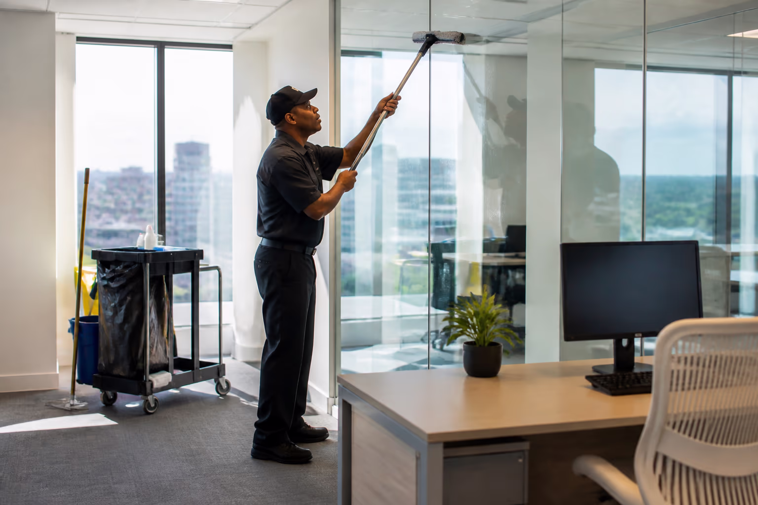 A janitorial technician in a dark uniform cleaning a glass office wall and desk beside a floor-care cart in a bright Ottawa office tower.