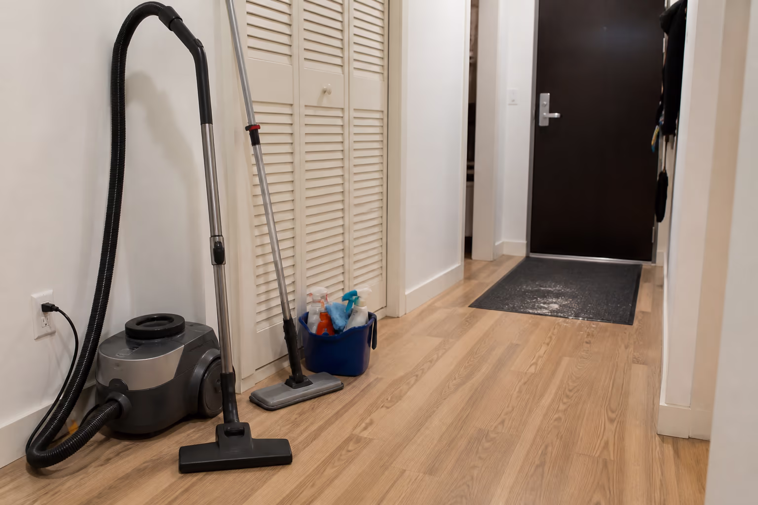 A landscape view of a small Ottawa condo hallway with a vacuum, a floor mop and a small bucket of cleaning supplies lined up beside a closet, with an entry mat catching grit at the door.