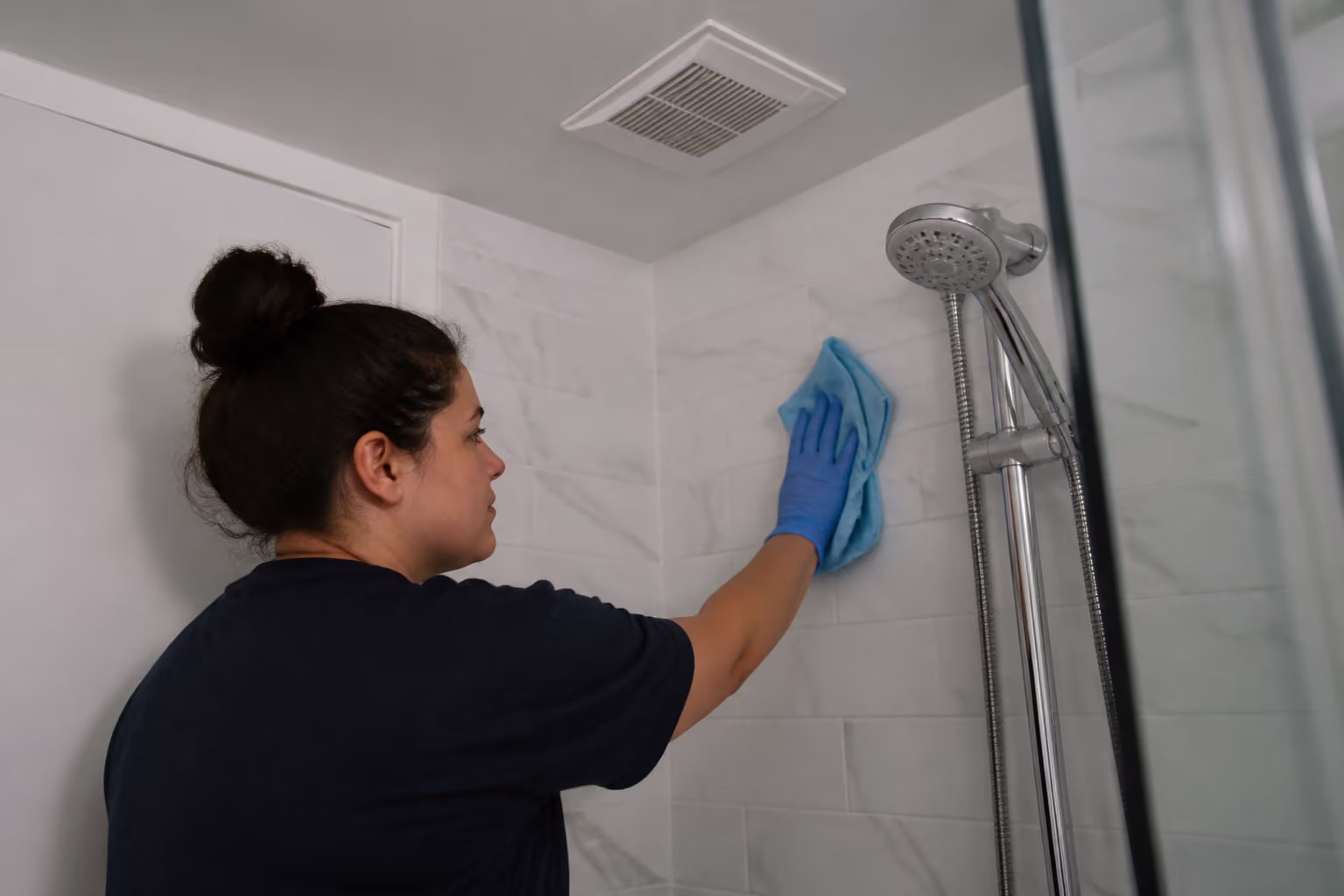 A wide view of a cleaner wiping a bathroom shower wall and grout with a microfibre cloth while an exhaust fan runs above, in a bright Ottawa bathroom.