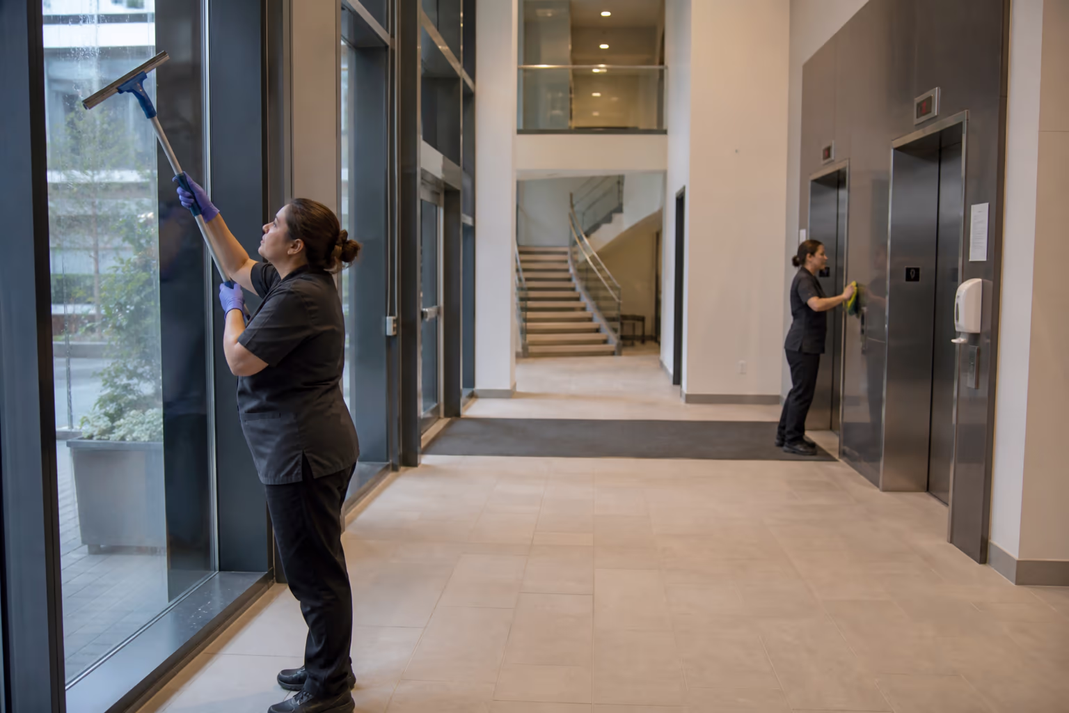 A uniformed cleaning team working a bright multi-floor Ottawa office lobby, one polishing entrance glass and another resetting a corridor, with elevators and a stairwell behind.