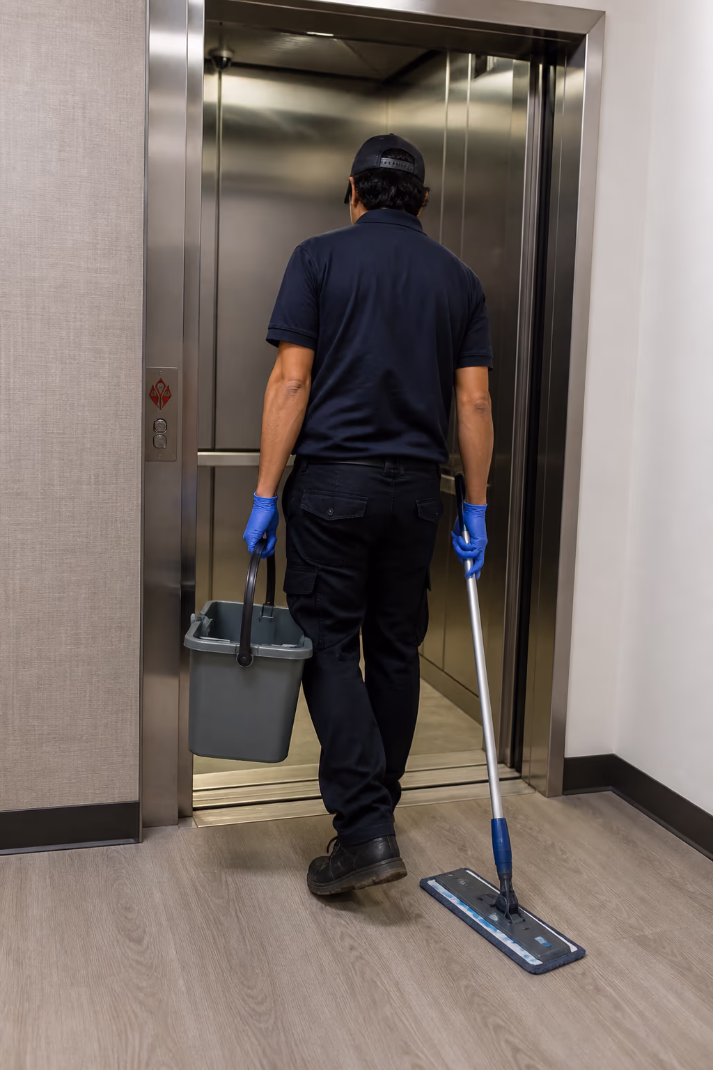 A portrait of a commercial cleaner carrying a microfibre mop and a closed bucket into a service elevator in an Ottawa tower, ready to work the upper floors at off-peak hours.