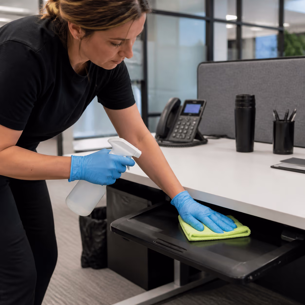 A close-up square view of a professional cleaner wiping a shared office desk and keyboard tray with a microfibre cloth and certified cleaning spray in a bright Ottawa workspace.