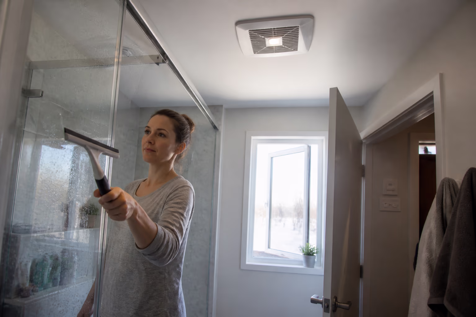 A bright Ottawa bathroom with a window cracked open and an exhaust fan running, a squeegee drying the shower glass, and the door left ajar to let winter moisture clear.