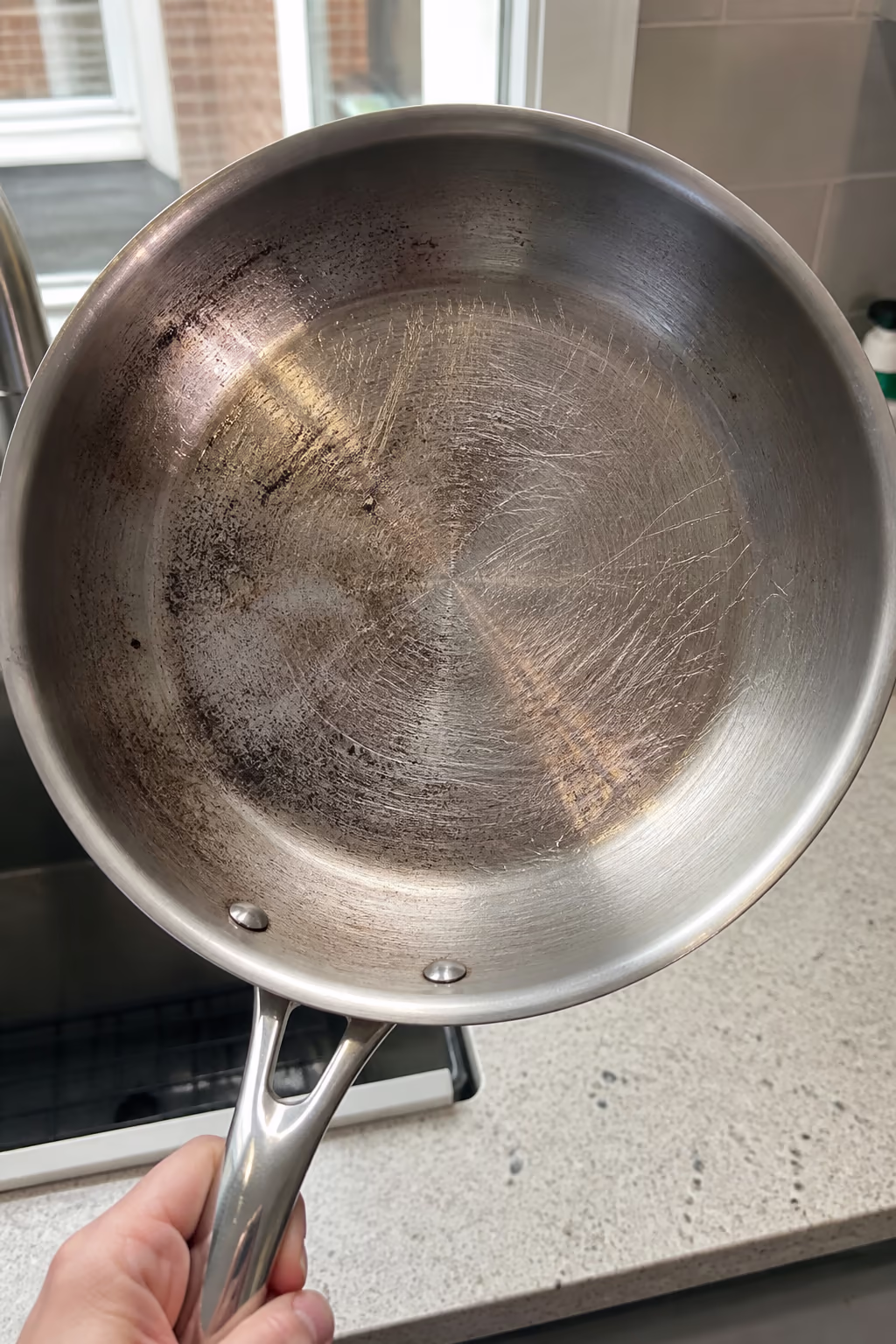 A portrait of a stainless steel pan with dull scratch marks across the interior from a metal scrubber, showing how steel wool permanently damages the finish and traps residue.