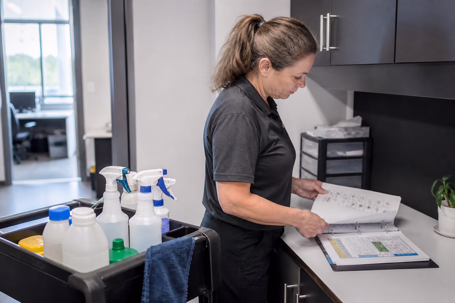 A landscape view of a professional office cleaning team reviewing a documented schedule and WHMIS safety data sheets at a workstation in an Ottawa office, with labelled cleaning products organized on a cart.