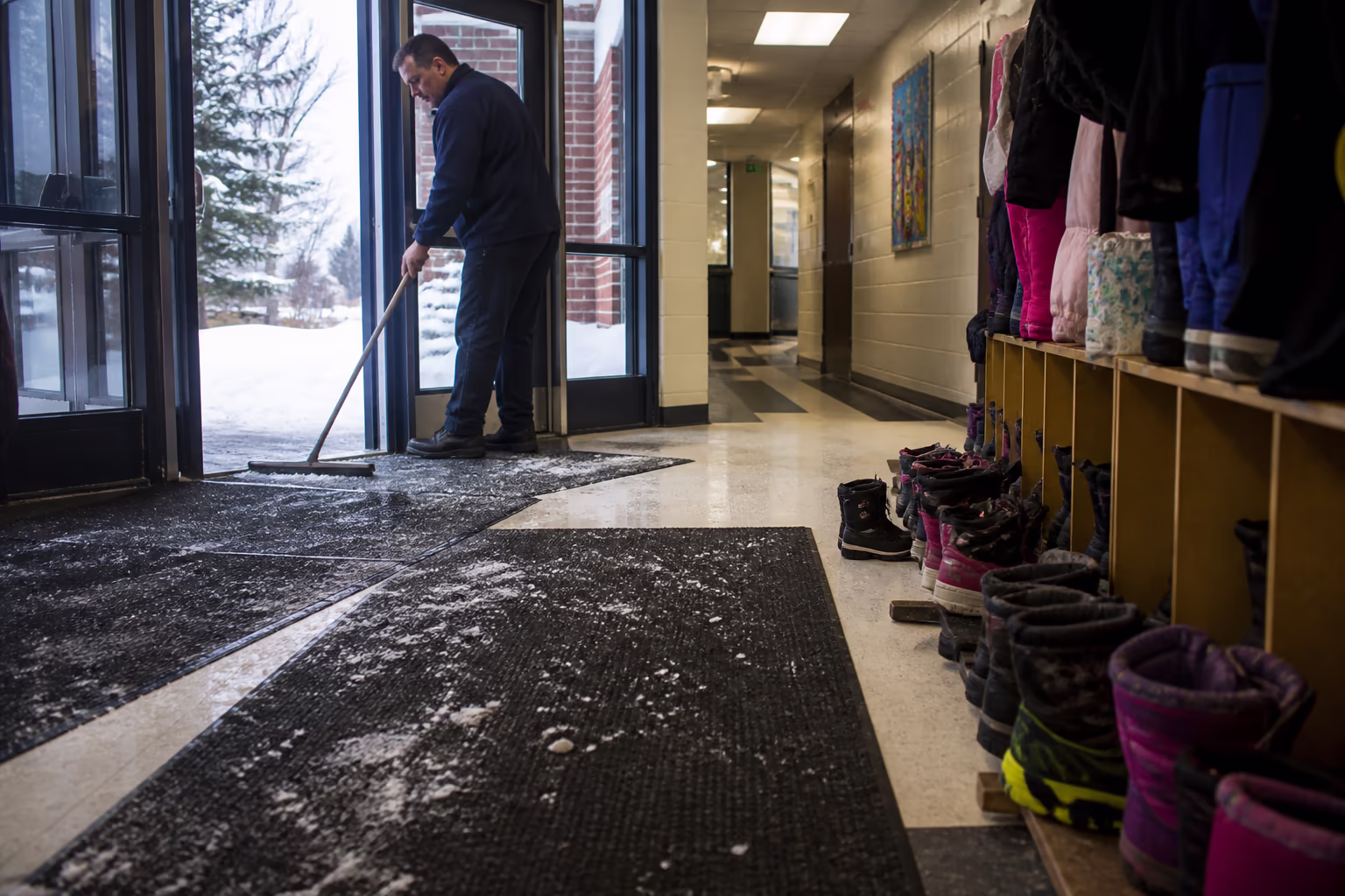 A wide view of a school entrance in winter, with entry mats laid out and a cleaner managing salt and slush at the door while boots line the hallway inside.