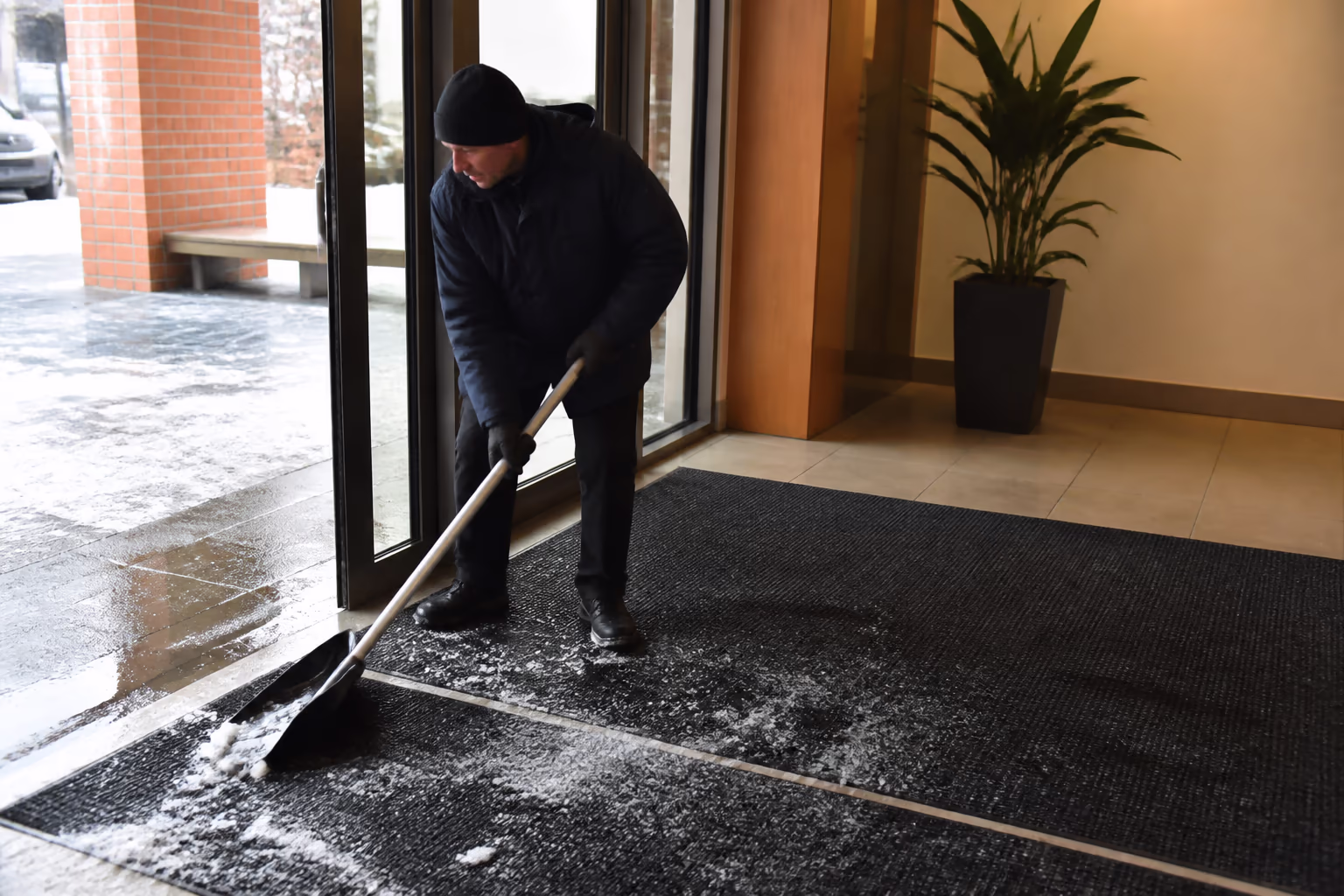 A wide view of an Ottawa office entrance in winter, with entry mats laid out and a cleaner managing salt and slush at the door while the lobby floor stays dry inside.