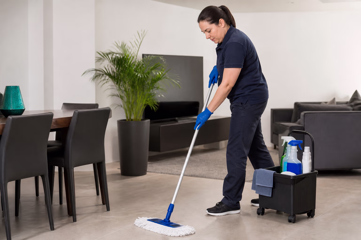 A uniformed janitorial team in a bright Ottawa building lobby during a staff training session, a supervisor demonstrating a cleaning technique to two new hires near the entrance and a supply cart.
