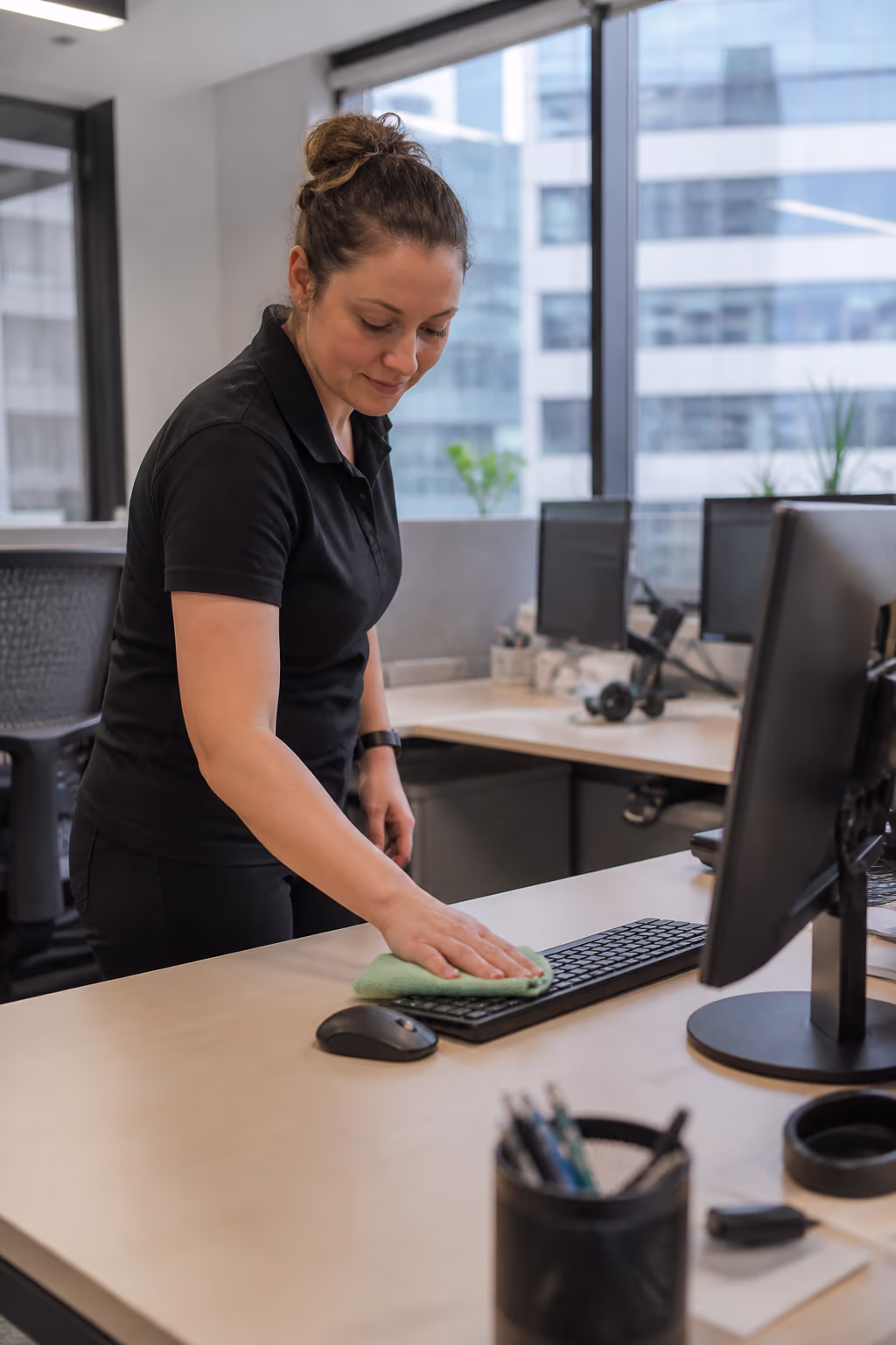 A portrait of a cleaner wiping a shared office desk and keyboard with a microfibre cloth during a daytime office cleaning pass in a bright Ottawa workspace.