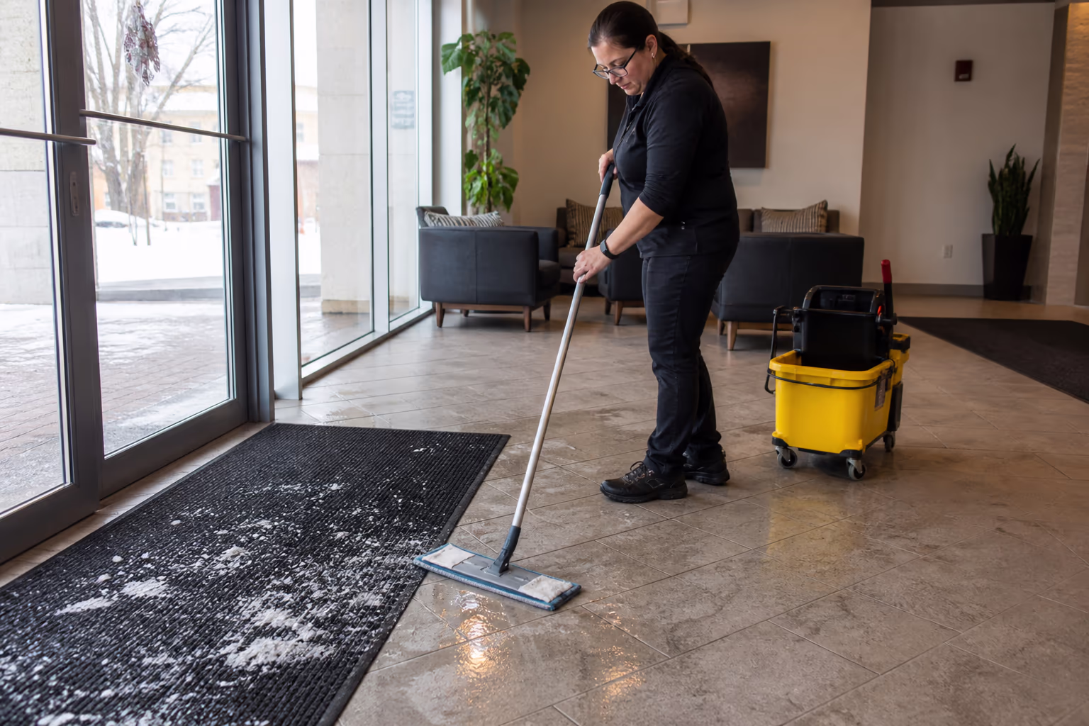 Entry matting and a salt-marked reception floor being swept and mopped by a uniformed commercial cleaner during a snowy Ottawa winter, with wet-day mats laid at the door.