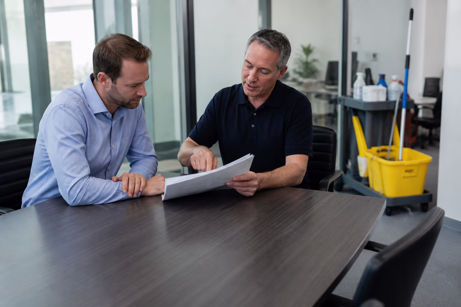 A janitorial supervisor walking a client through an itemised cleaning contract and pricing breakdown at a conference table in a bright Ottawa office, with a mop bucket and supply cart nearby.