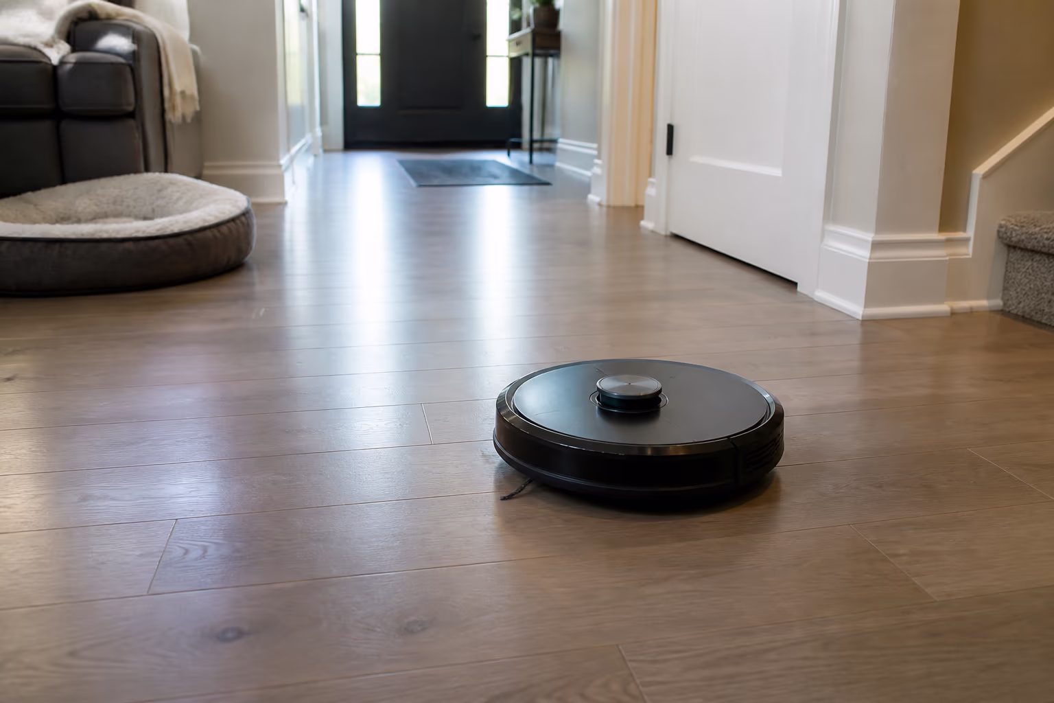 A robotic vacuum gliding across the sealed hard-floor entryway of an Ottawa home, its sensors mapping the room while a sofa and pet bed stay out of the way, capturing the hands-free daily maintenance a robot adds to a cleaning routine.