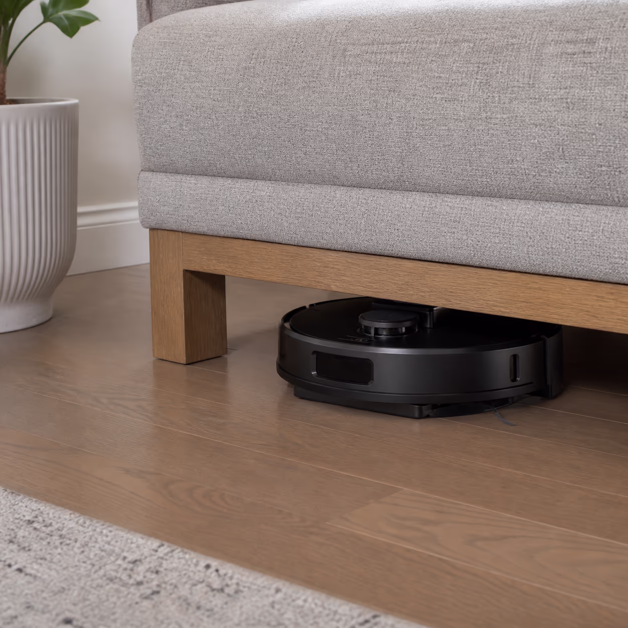 A square view of a robotic vacuum parked in its charging dock under a low sofa in an Ottawa living room, capturing how the robot slides into the everyday layer of a home cleaning routine.