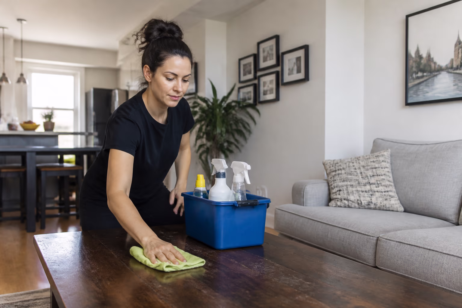 A portrait of a residential cleaner in a bright Ottawa living room setting out supplies for a first visit, wiping a high-traffic surface and noting the deeper work for the plan, capturing the measured, honest start to a home cleaning routine.