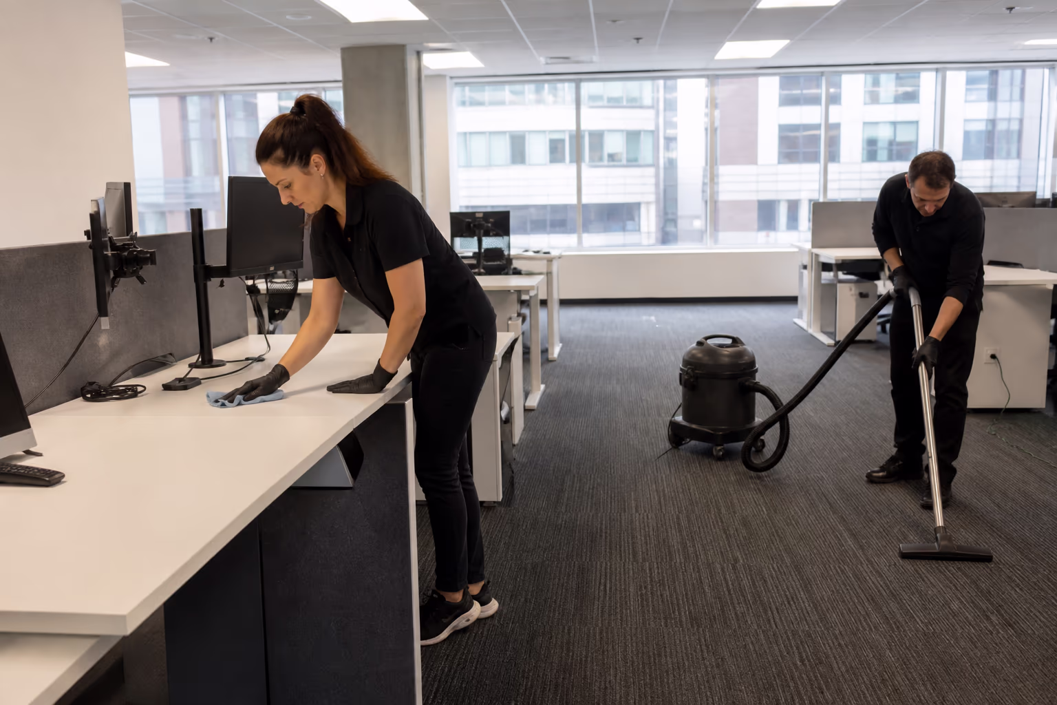 A portrait of a professional cleaning team working an open-plan Ottawa office, one cleaner wiping a shared desk while a second vacuums the aisle, capturing the everyday build-up that signals the routine is not keeping up.