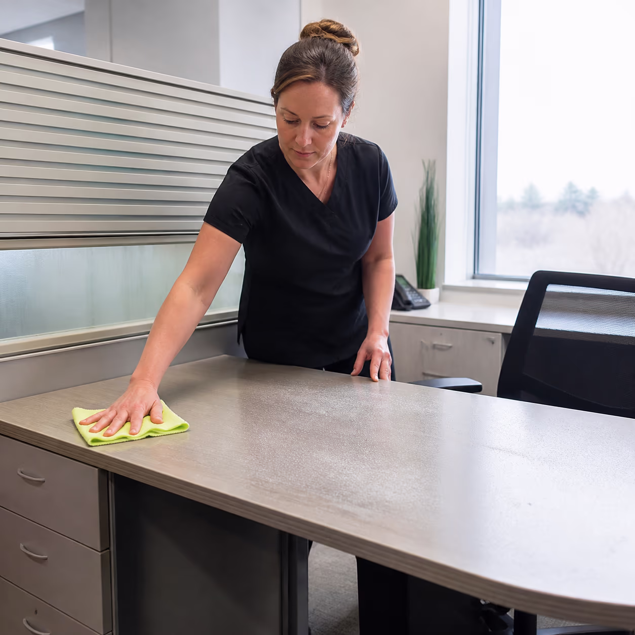A square view of a cleaner dusting a shared office desk and partition in an Ottawa workspace, showing the quiet build-up that collects when a routine is not keeping up.
