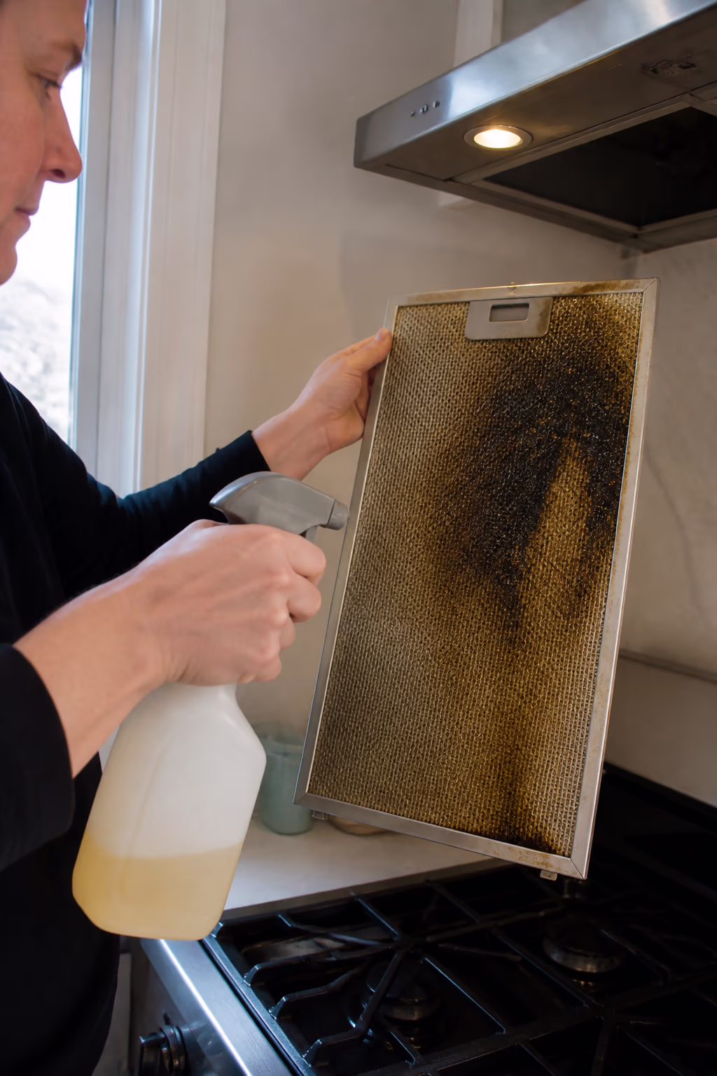 A portrait of a cleaner applying a degreaser to a greasy range hood filter in an Ottawa kitchen, showing the heavy baked-on grease that soap alone cannot lift.