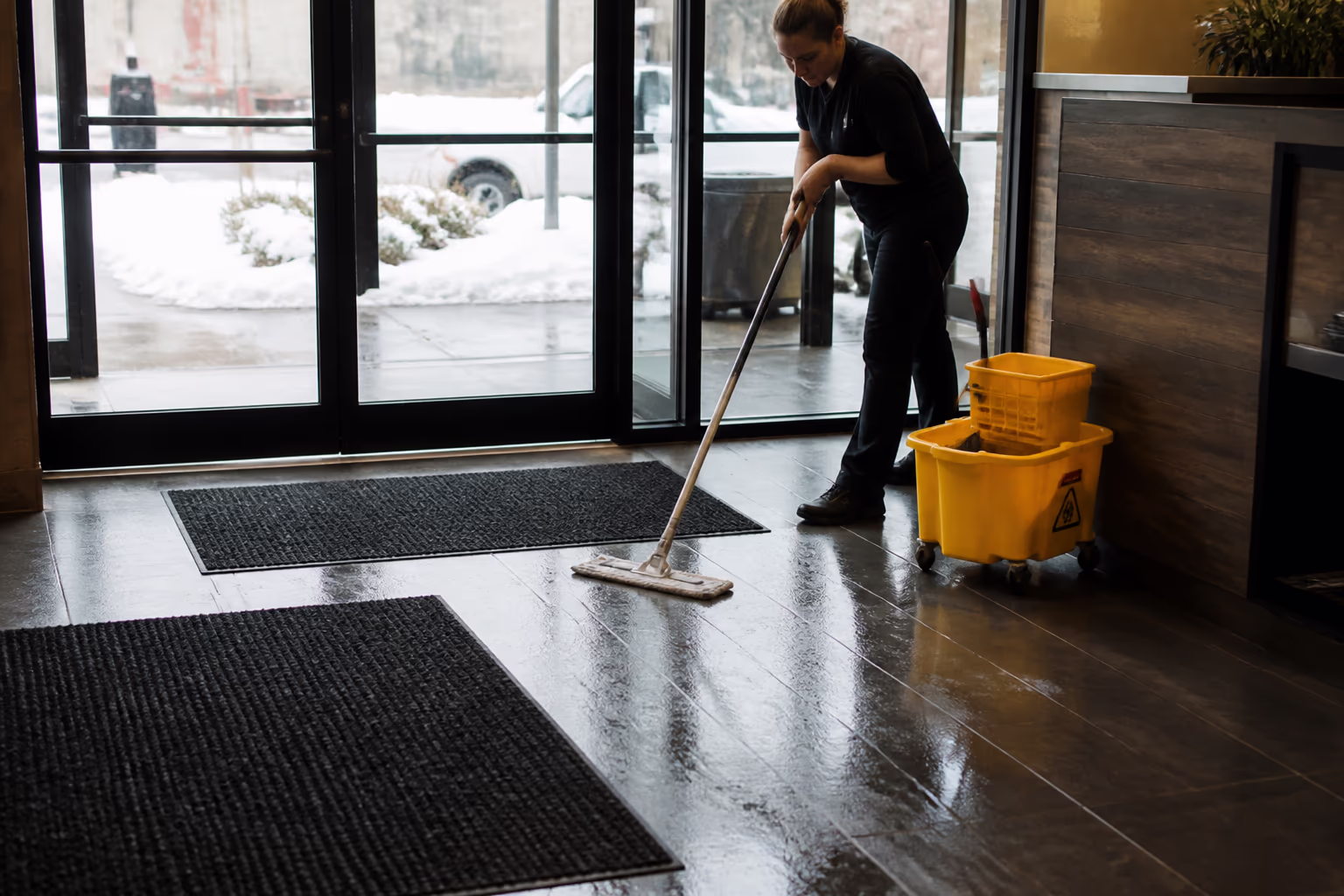 A wide view of an Ottawa restaurant entrance in winter with entry mats laid out and a staff member mopping the salt and slush that are tracked in, capturing the seasonal routine that keeps floors clean and safe.