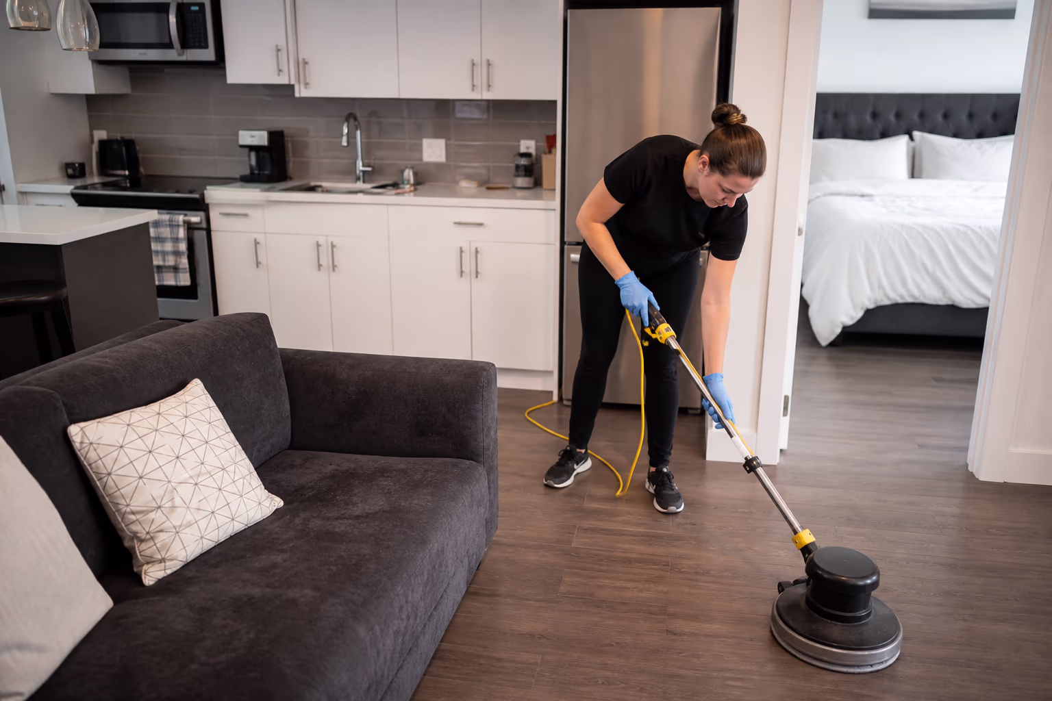 A residential cleaner turning over a compact short-term rental unit in Ottawa, stripping the bed and wiping the kitchen counters between guest stays, capturing the tight turnaround work at the heart of Airbnb cleaning.