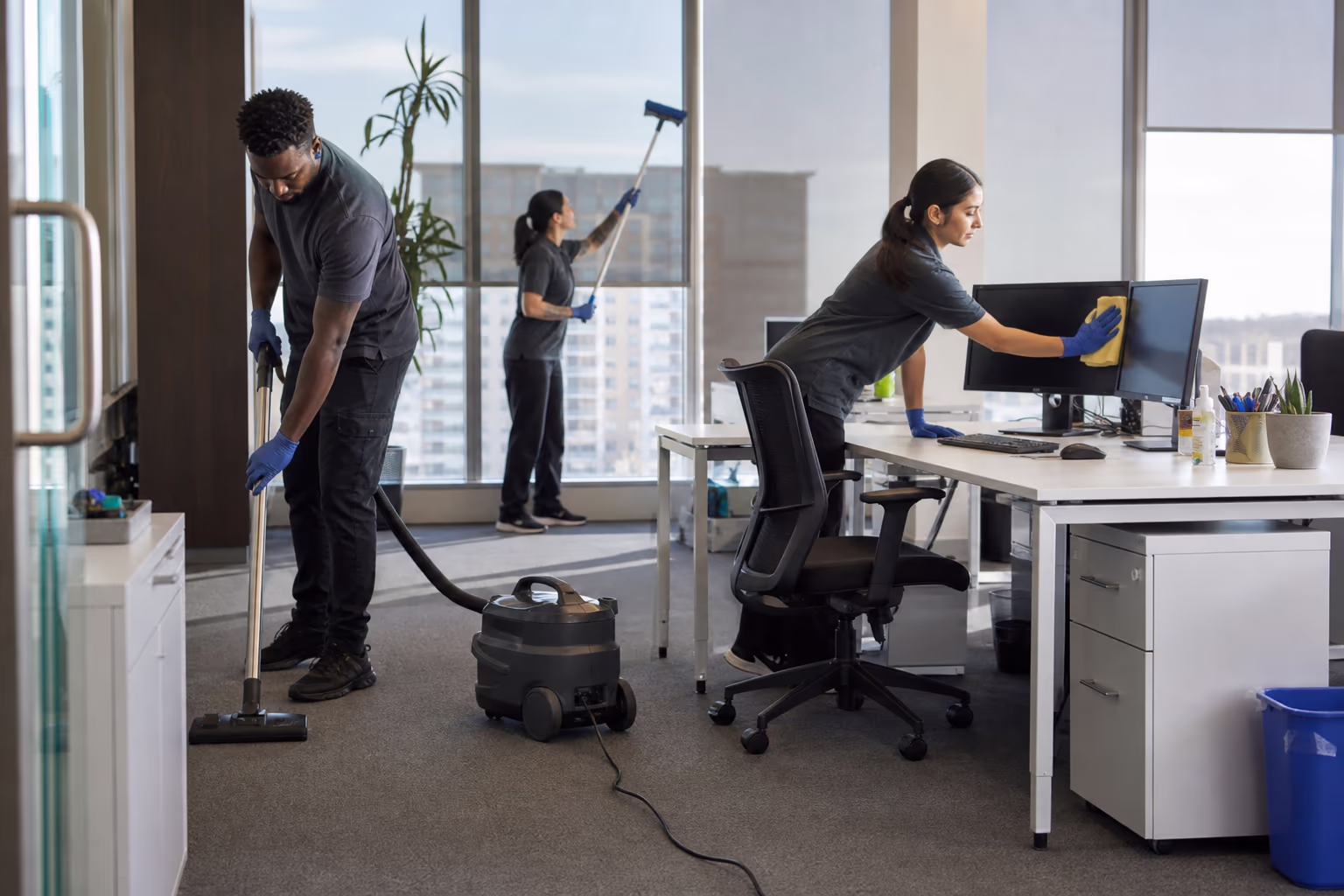 Three cleaners in an office: one vacuuming the floor, one wiping a monitor, one washing a window with a pole
