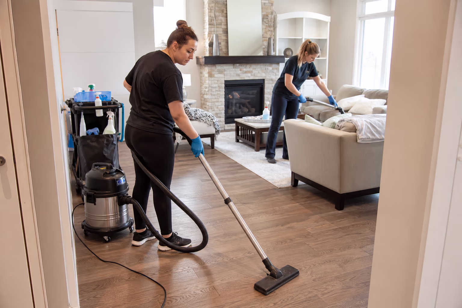 Two cleaners vacuuming a living room beside a cleaning cart