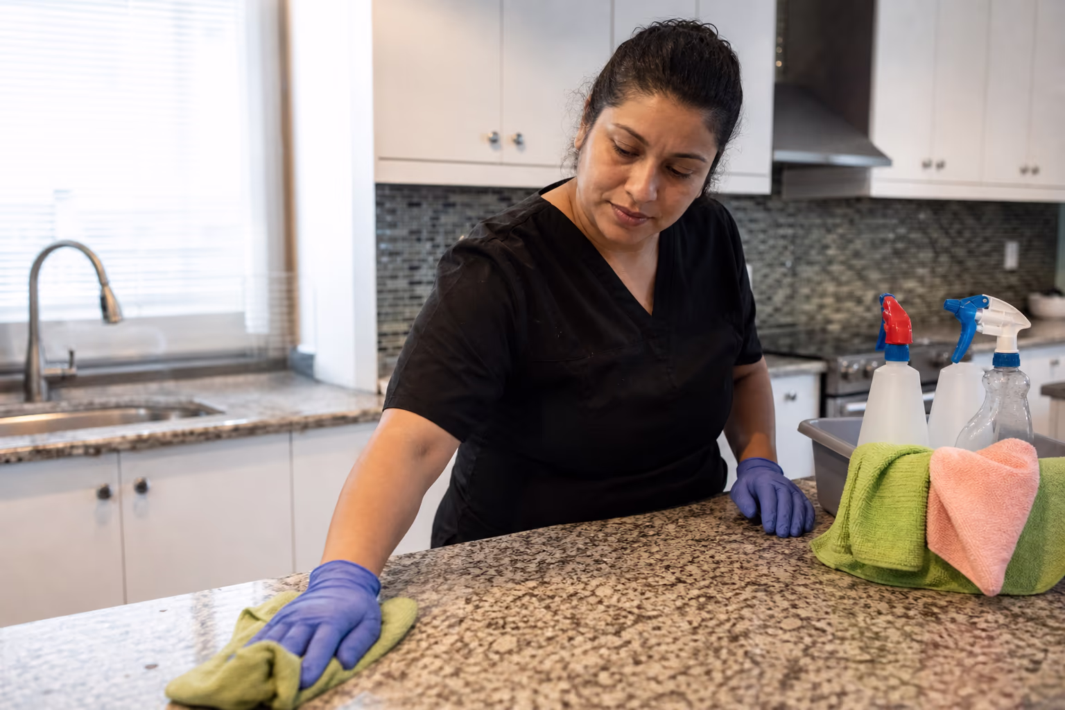 A cleaner wiping a kitchen counter beside folded cloths and spray bottles in a bright home kitchen.