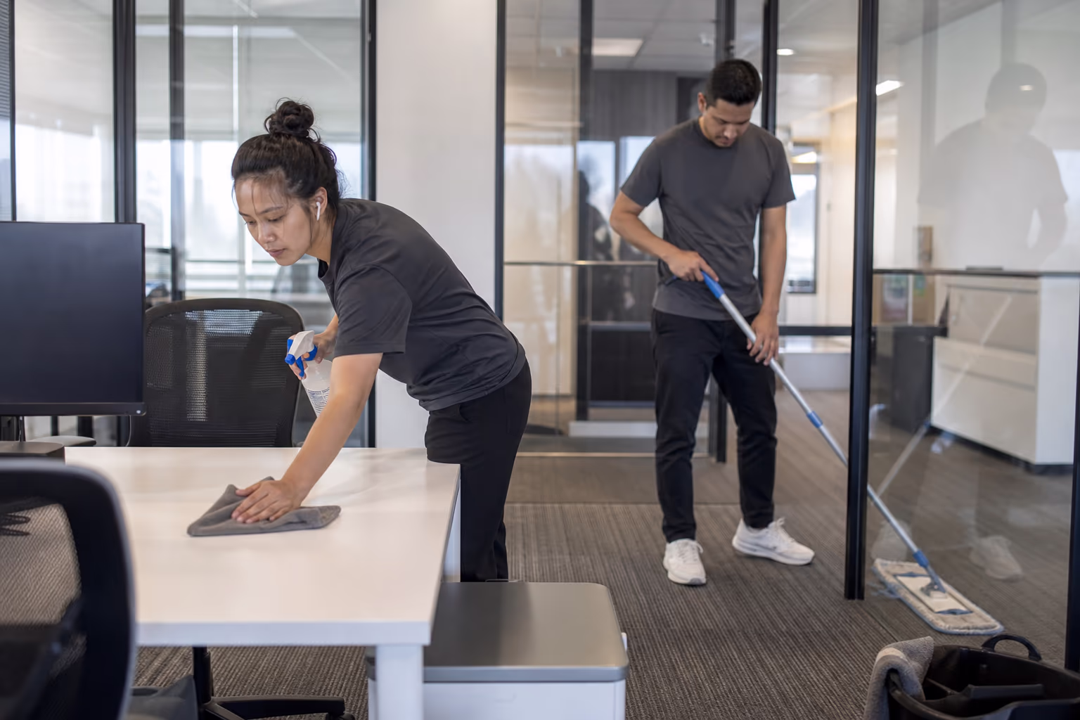 Two cleaners wiping a desk and mopping a floor in a glass-walled office