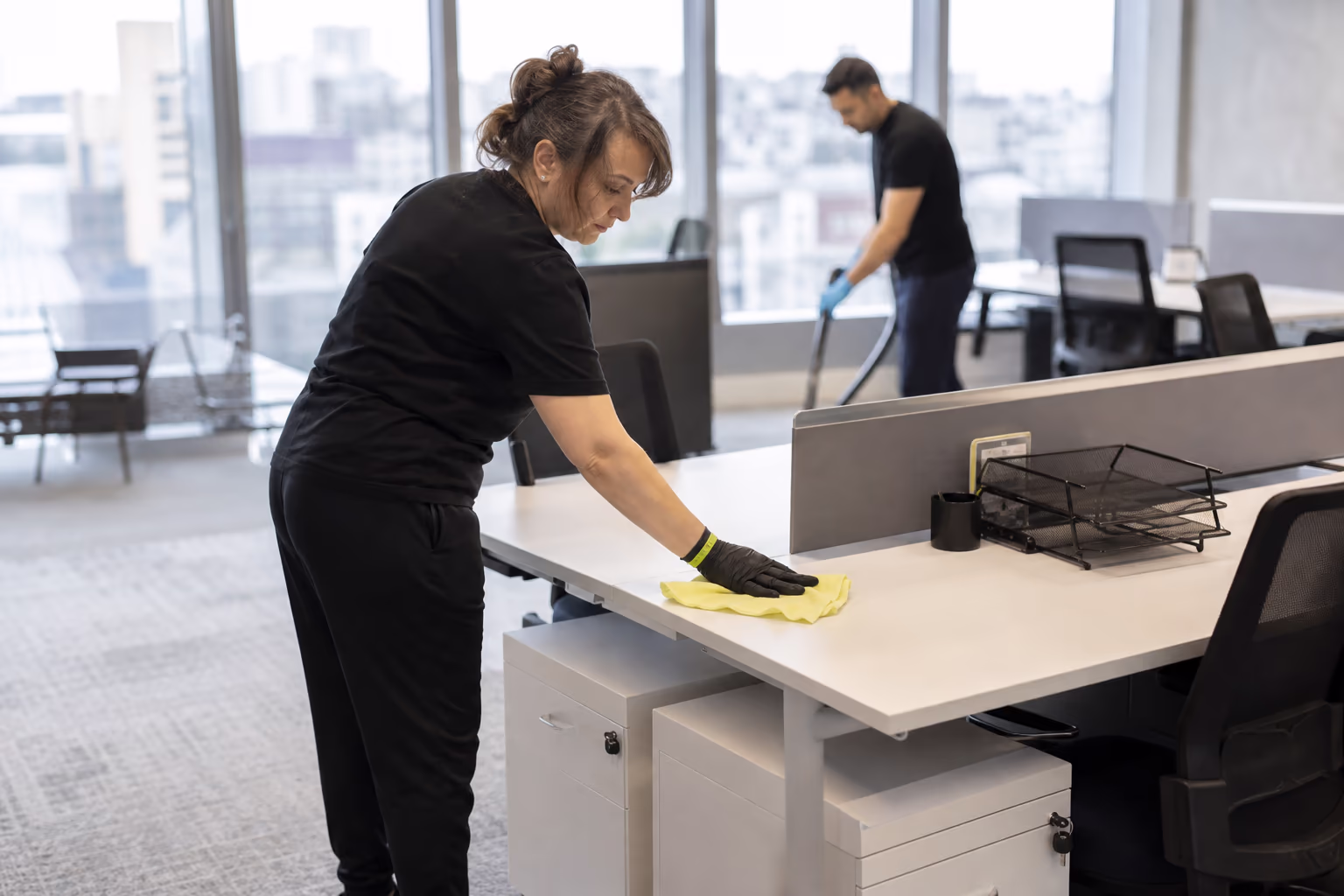 Cleaners in a bright office, one dusting a desk while another works the open floor