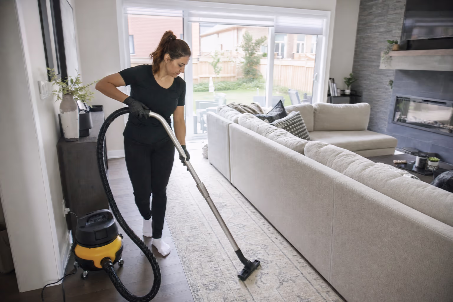 Cleaner vacuuming a bright living room beside a wall of daylight windows